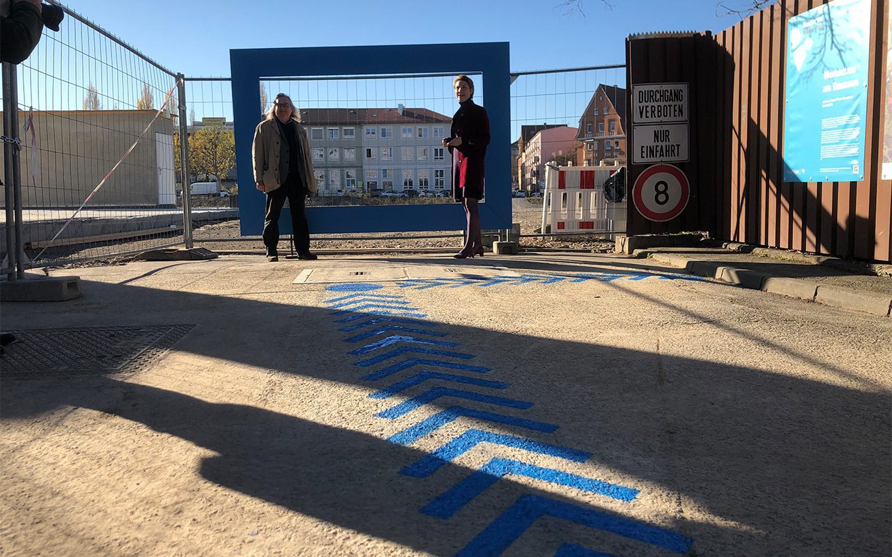In Form von Pfeilen und Kreis hat Patrick ein Blaues Band für die Wegführung zur Werkschau am Bauzaun des Neubauprojekts “Quartier am Wiener Platz“ gesprüht.