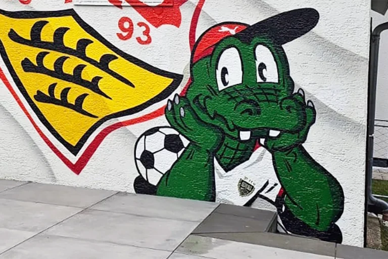 Eine weiß gestrichene Garagenwand mit einem Graffiti des VfB Stuttgart-Wappens und dem grün lächelnden Maskottchen Fritzle, der einen Fußball hält