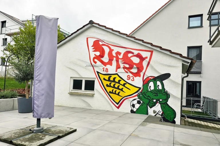 Eine weiß gestrichene Garagenwand mit einem Graffiti des VfB Stuttgart-Wappens und dem grün lächelnden Maskottchen Fritzle, der einen Fußball hält